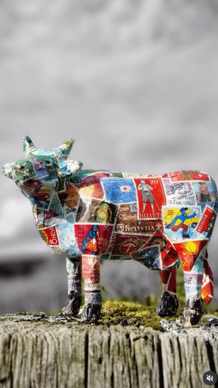 Muh — Kuh-Objekt, beklebt mit Schweizer Briefmarken, auf einem Holzstamm vor Bergkulisse